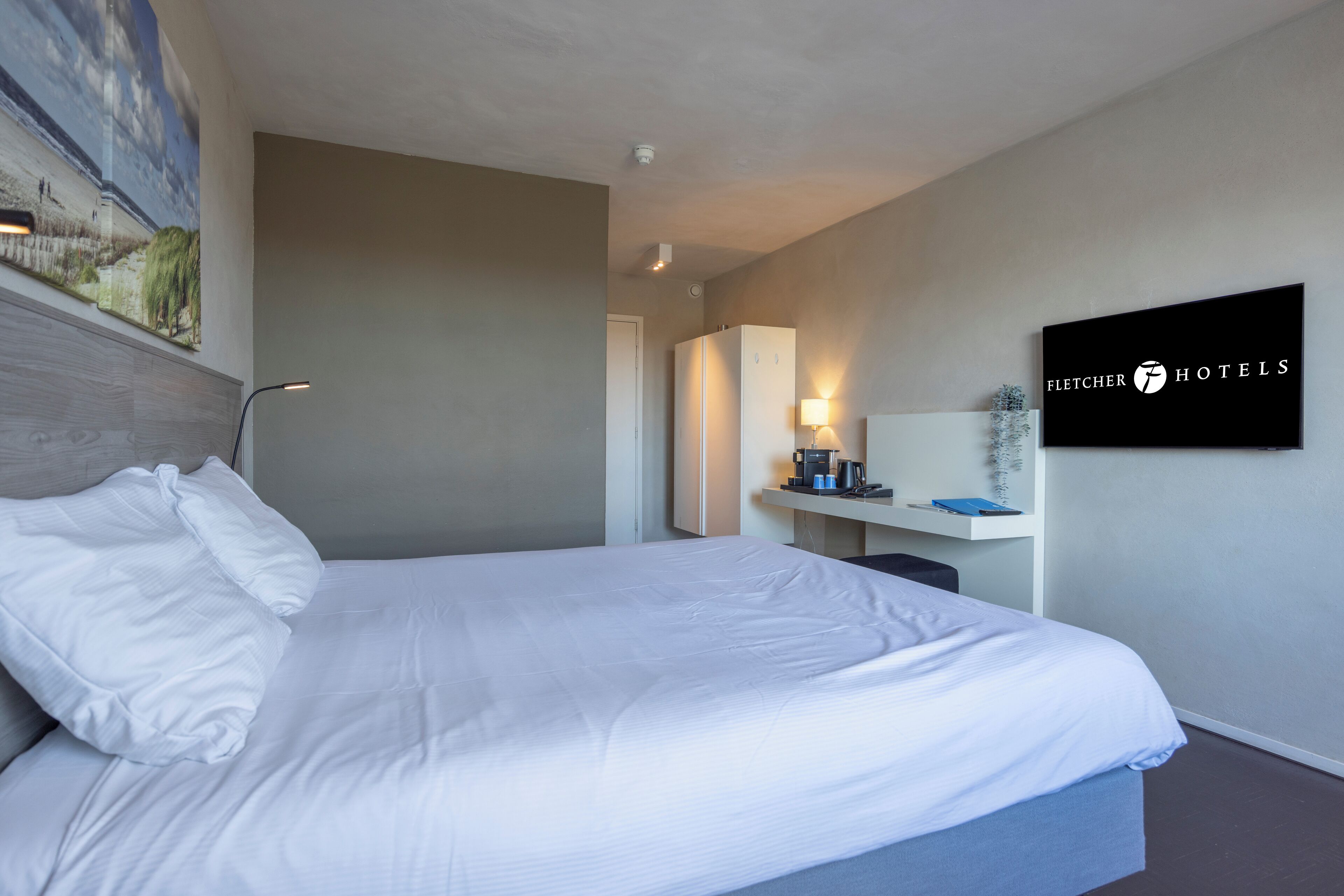 Una habitación de hotel con una cama, un escritorio, una silla, una lámpara, una cafetera y un cuadro en la pared.