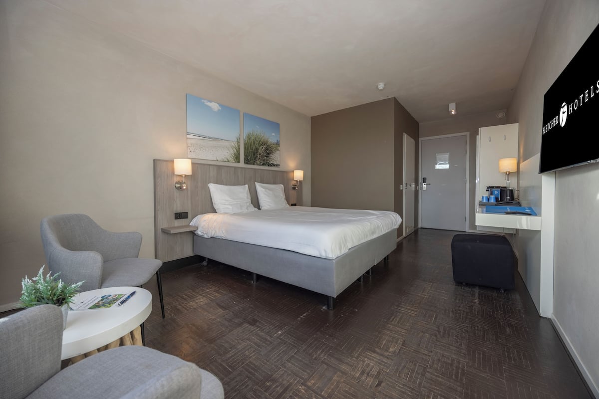 Quarto de hotel com uma cama, duas mesas de cabeceira com luminárias, uma pequena mesa redonda com uma planta, uma poltrona cinza, uma escrivaninha com um computador e uma televisão na parede.