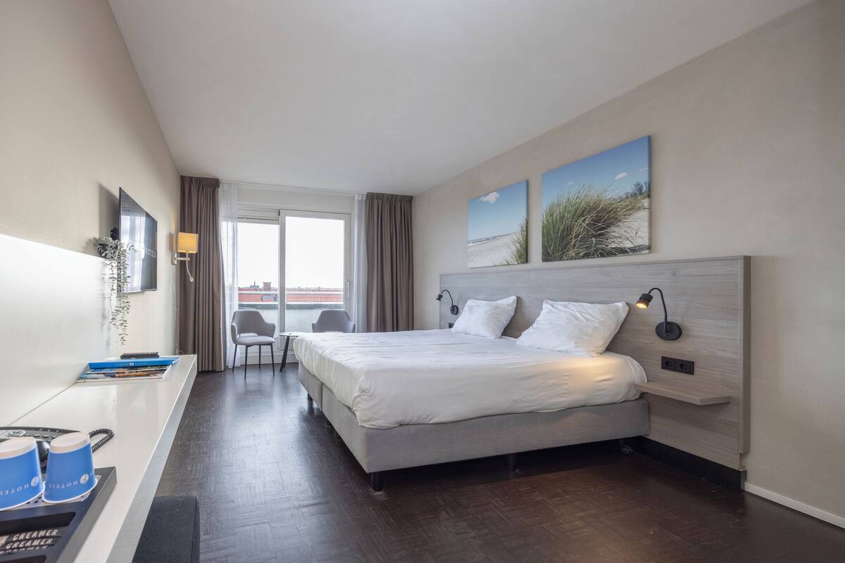 Quarto de hotel moderno com uma cama grande, uma escrivaninha com computador e vista para a cidade.
