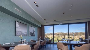 Un comedor con mesas y sillas redondas, ventanas amplias con vistas al campo y obras de arte en las paredes.