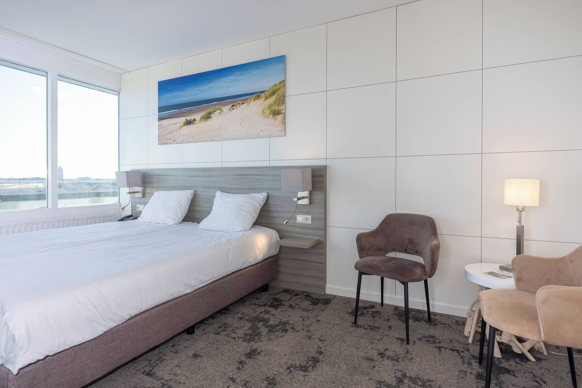 Quarto de hotel com uma cama grande, cabeceira com pintura de uma praia, mesa redonda pequena com abajur e duas cadeiras.