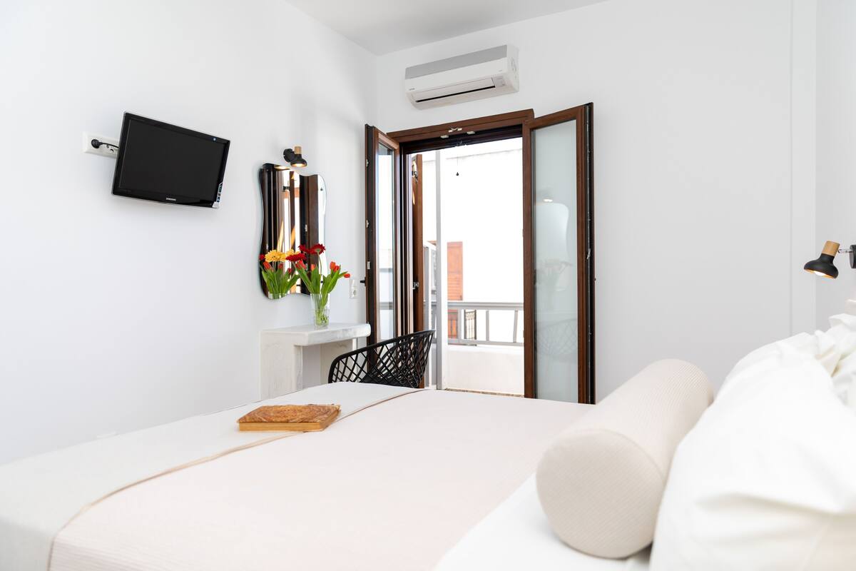 A modern bedroom with a bed, a small table with a book, a vase with flowers, and a TV mounted on the wall.