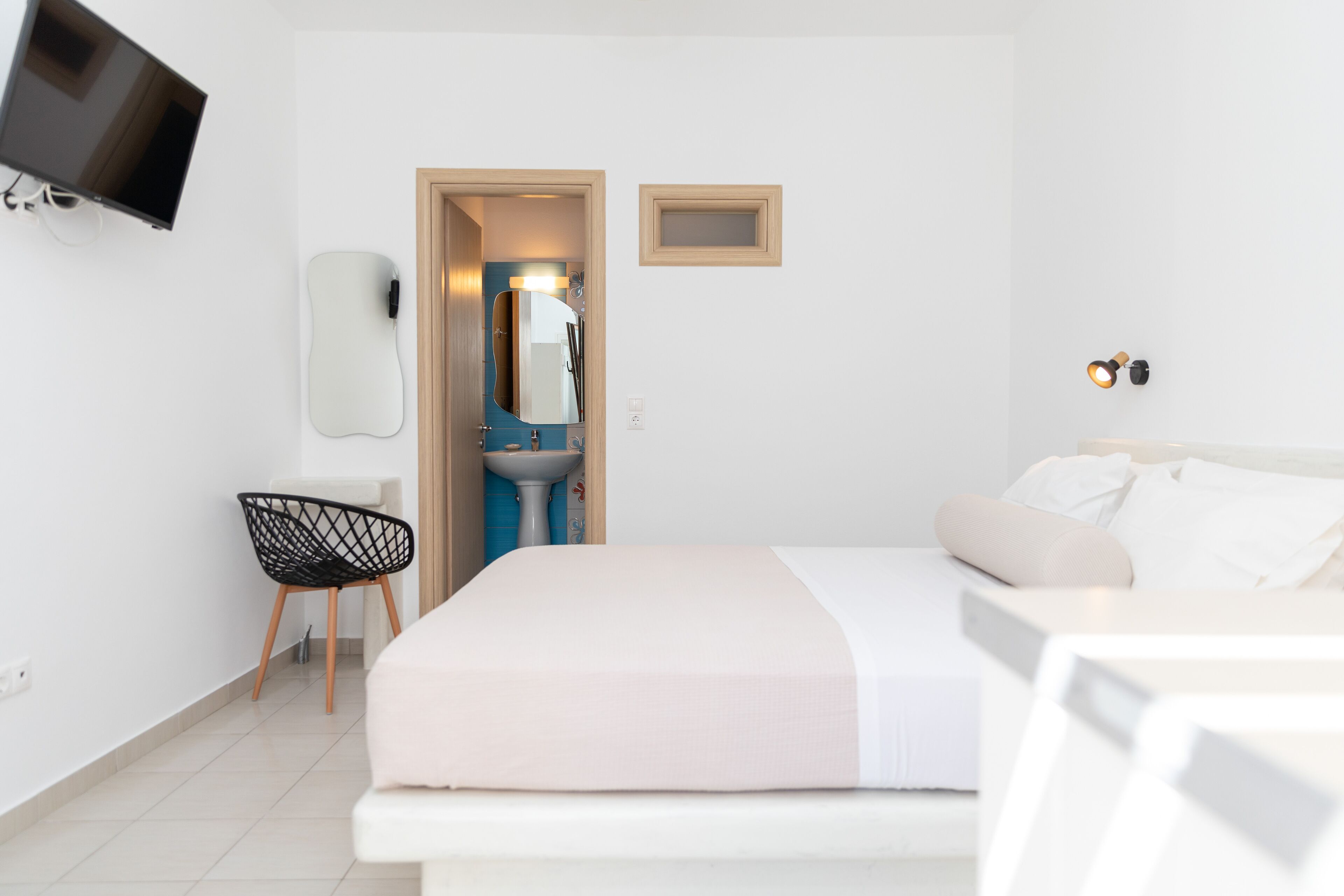 A modern hotel room with a bed, a TV mounted on the wall, a small desk with a chair, and a bathroom visible through an open door.
