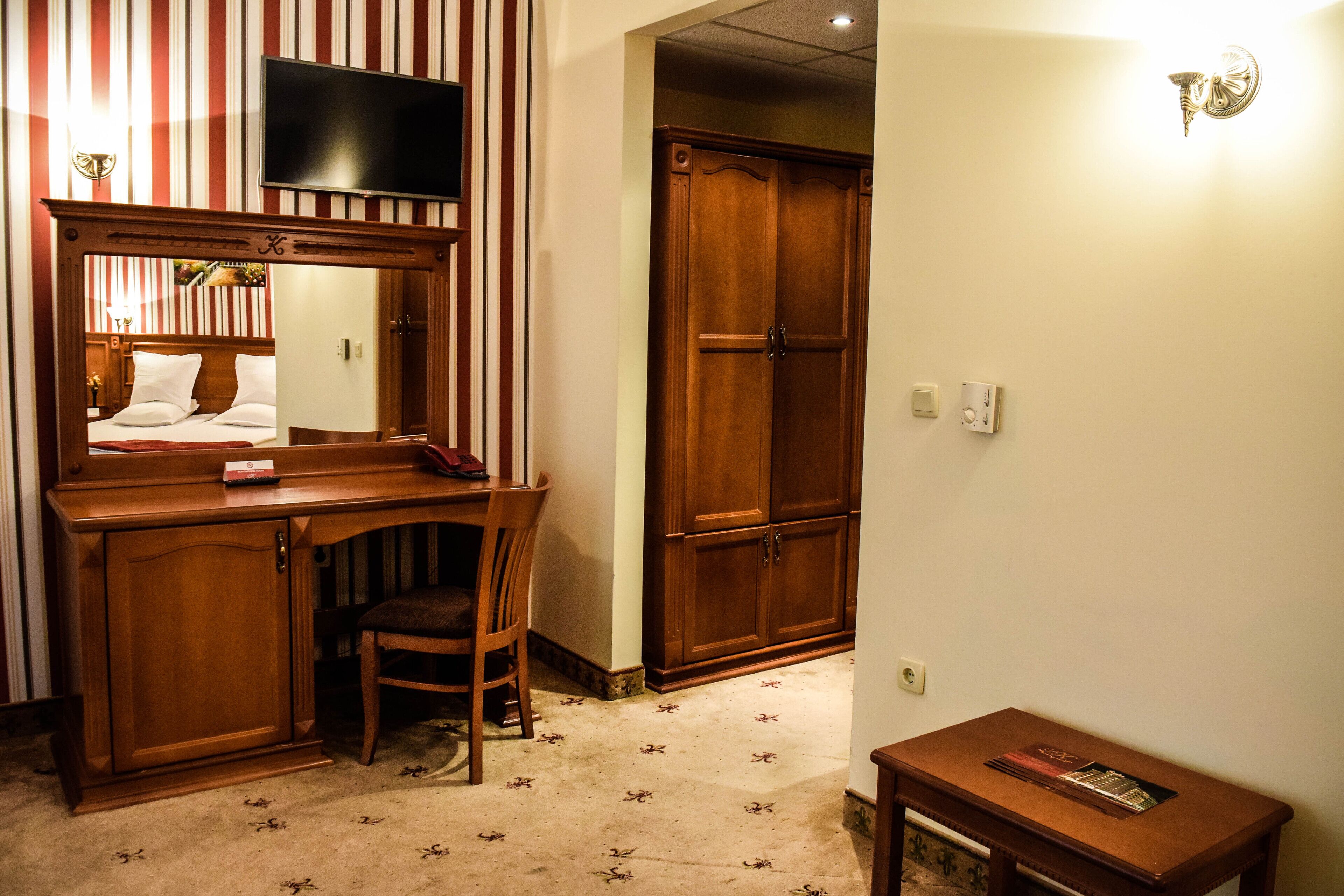 A hotel room with a wooden desk, a chair, a television mounted above the desk, a wardrobe, and a small table with a book.