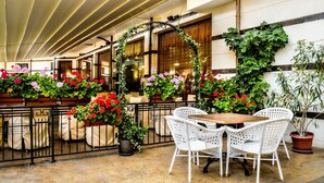 An outdoor seating area with white wicker chairs and tables, surrounded by potted plants and flowers. - Hotel Karolina (Sunny Beach)