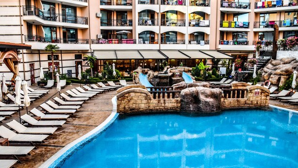 A swimming pool with lounge chairs and a water slide, surrounded by a multi-story hotel building with balconies. - Hotel Karolina (Sunny Beach)