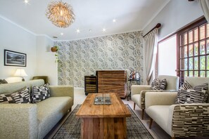 A modern living room with two sofas, a wooden coffee table, patterned wallpaper, and a large window with curtains. - Hlangana Lodge (Oudtshoorn)
