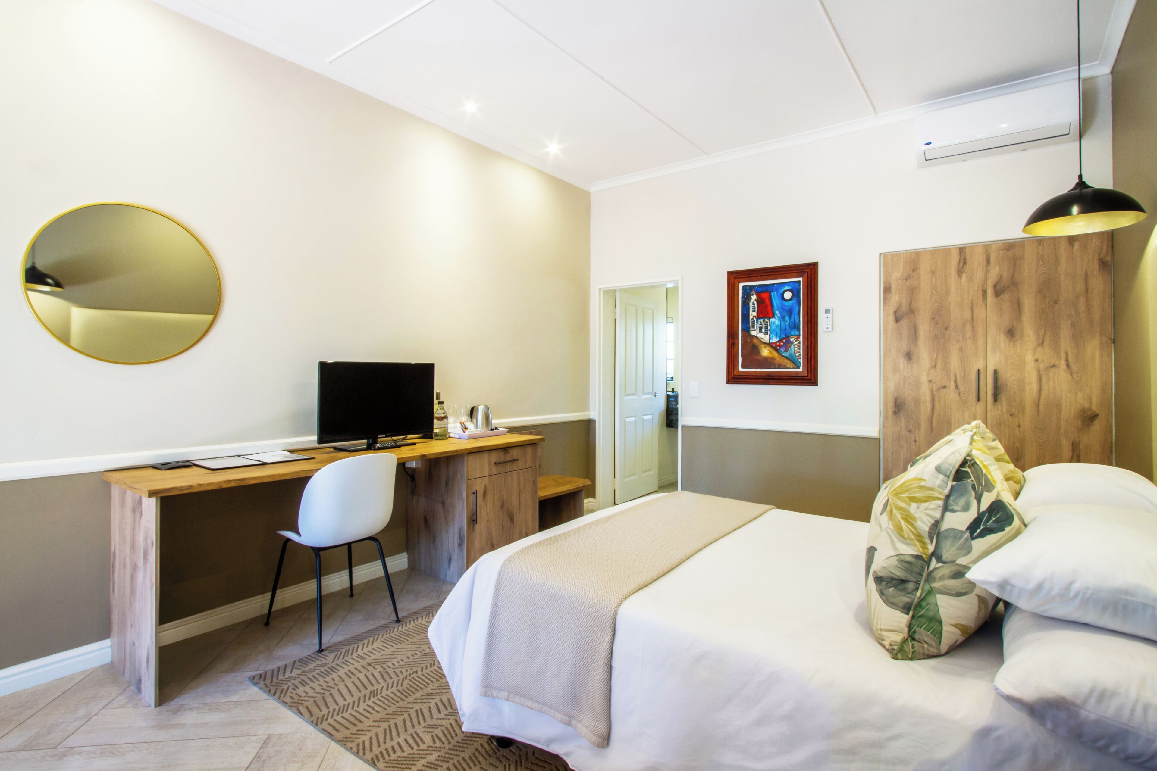 Una habitación de hotel con una cama, un escritorio con computadora, una silla, un espejo redondo y un cuadro en la pared.