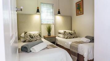 A hotel room with two beds, a window with blinds, a bedside table with a plant, and a painting on the wall.