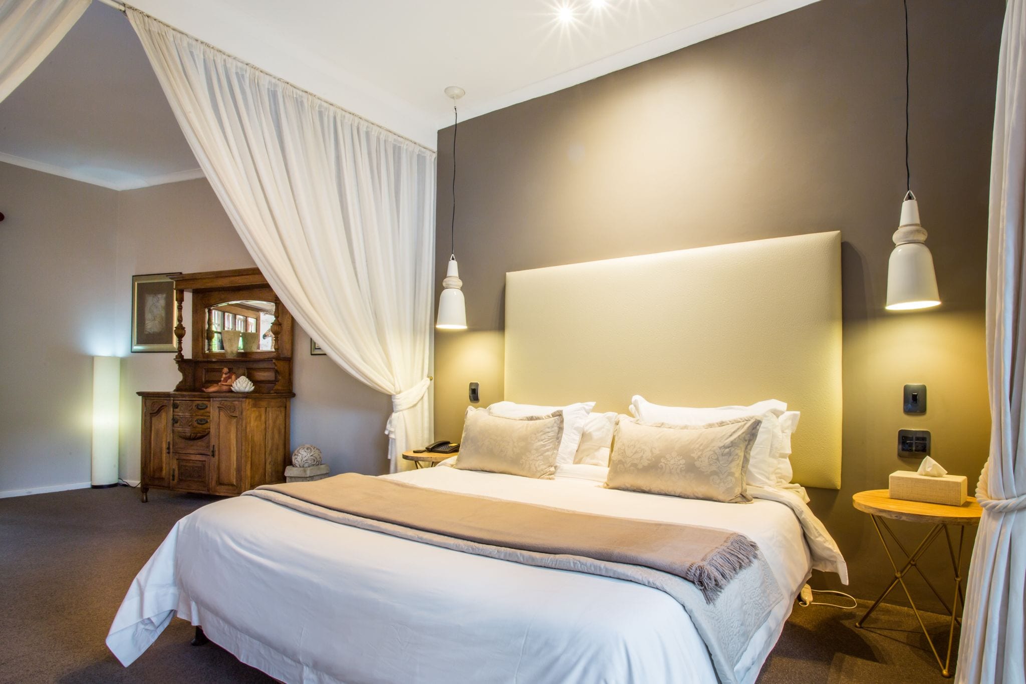 Una cama bien hecha con un cabecero grande, dos lámparas de noche, una mesita de noche de madera con una lámpara y un espejo en la pared.