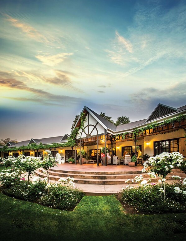 A hotel with a covered outdoor area, steps leading to a patio, and a garden with white flowers. - Hlangana Lodge (Oudtshoorn)