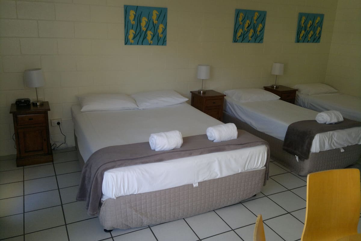 A hotel room with three beds, each with white linens and towels. There are two wall-mounted lamps on wooden nightstands.