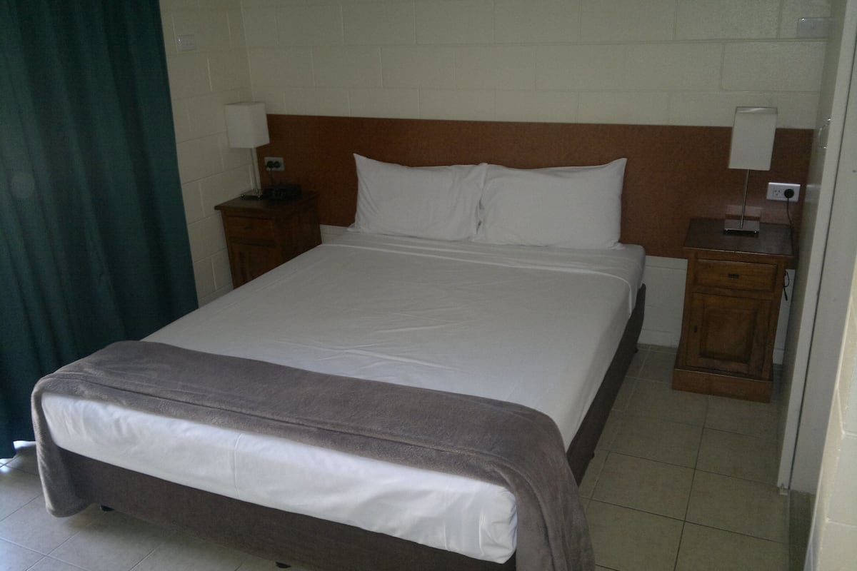 A standard hotel room with a single bed, wooden headboard, two bedside tables with lamps, and a green curtain.