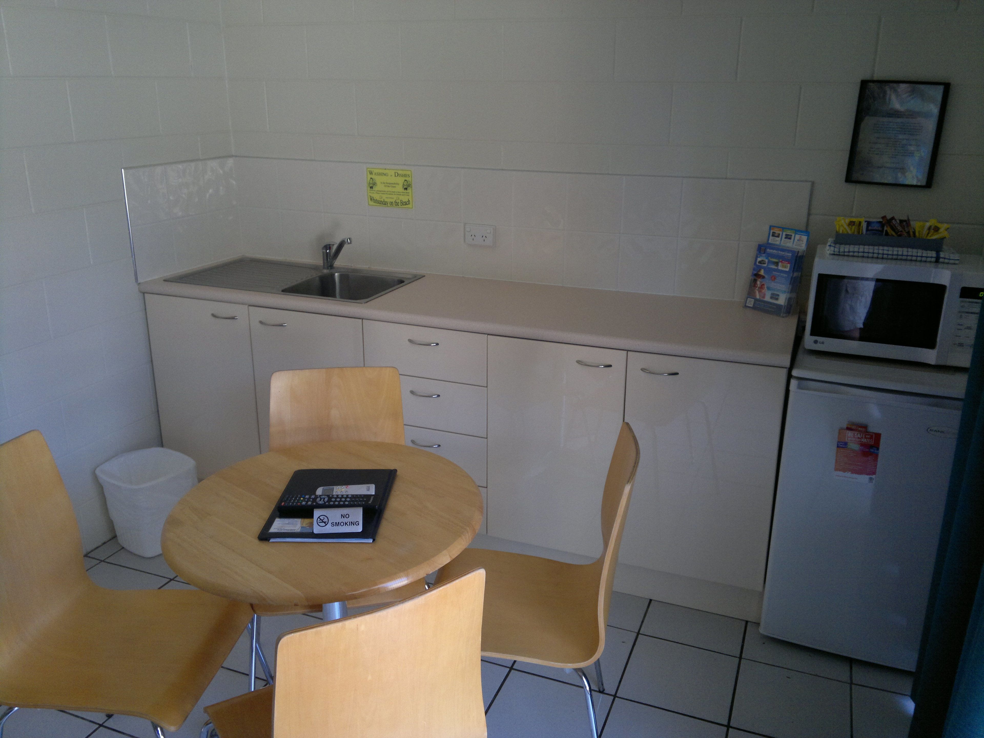 A small kitchenette with a sink, microwave, and refrigerator. There is a table with two chairs and a sign prohibiting smoking.