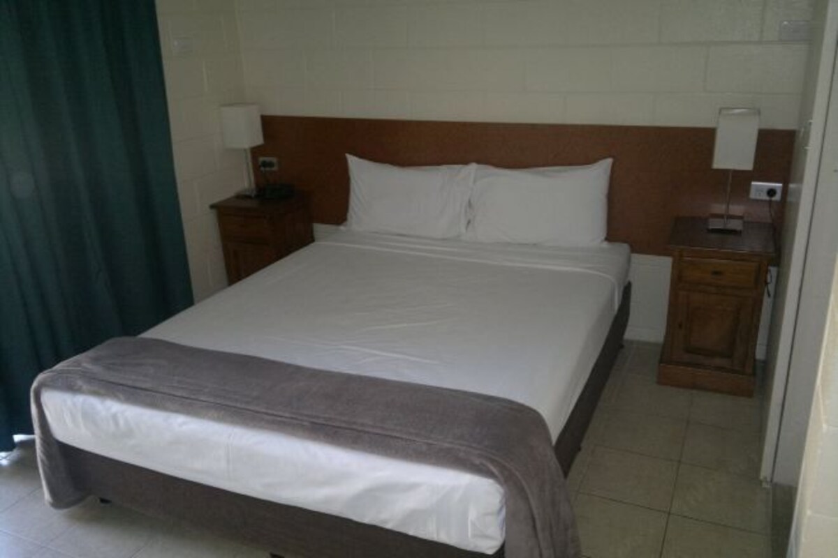 A hotel room with a large bed, wooden headboard, two bedside tables with lamps, and a curtain on one side.