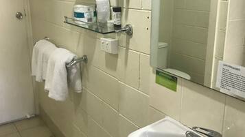 Un bagno con un lavandino bianco, uno specchio e un portasciugamani con un asciugamano bianco. Sono presenti articoli da toilette su uno scaffale sopra il lavandino.