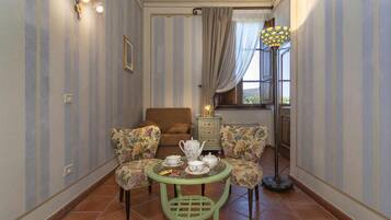 Ein Zimmer mit Streifenmuster-Tapete, einem braunen Sofa, zwei floralen Sesseln, einem grünen Tisch mit Teekanne, Tassen und Untertassen, einer Stehlampe und einem Fenster mit Vorhängen.