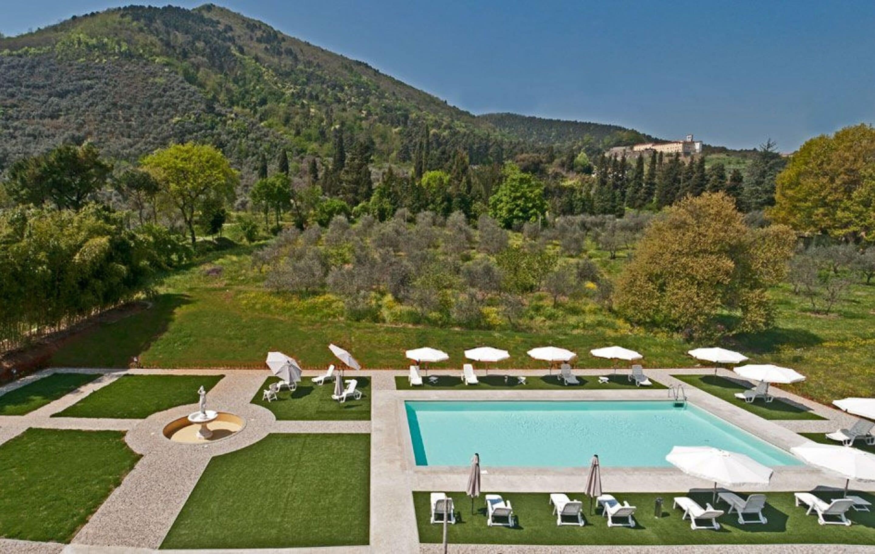 Zona de piscina con tumbonas y sombrillas, rodeada de césped cuidado y un paisaje montañoso.