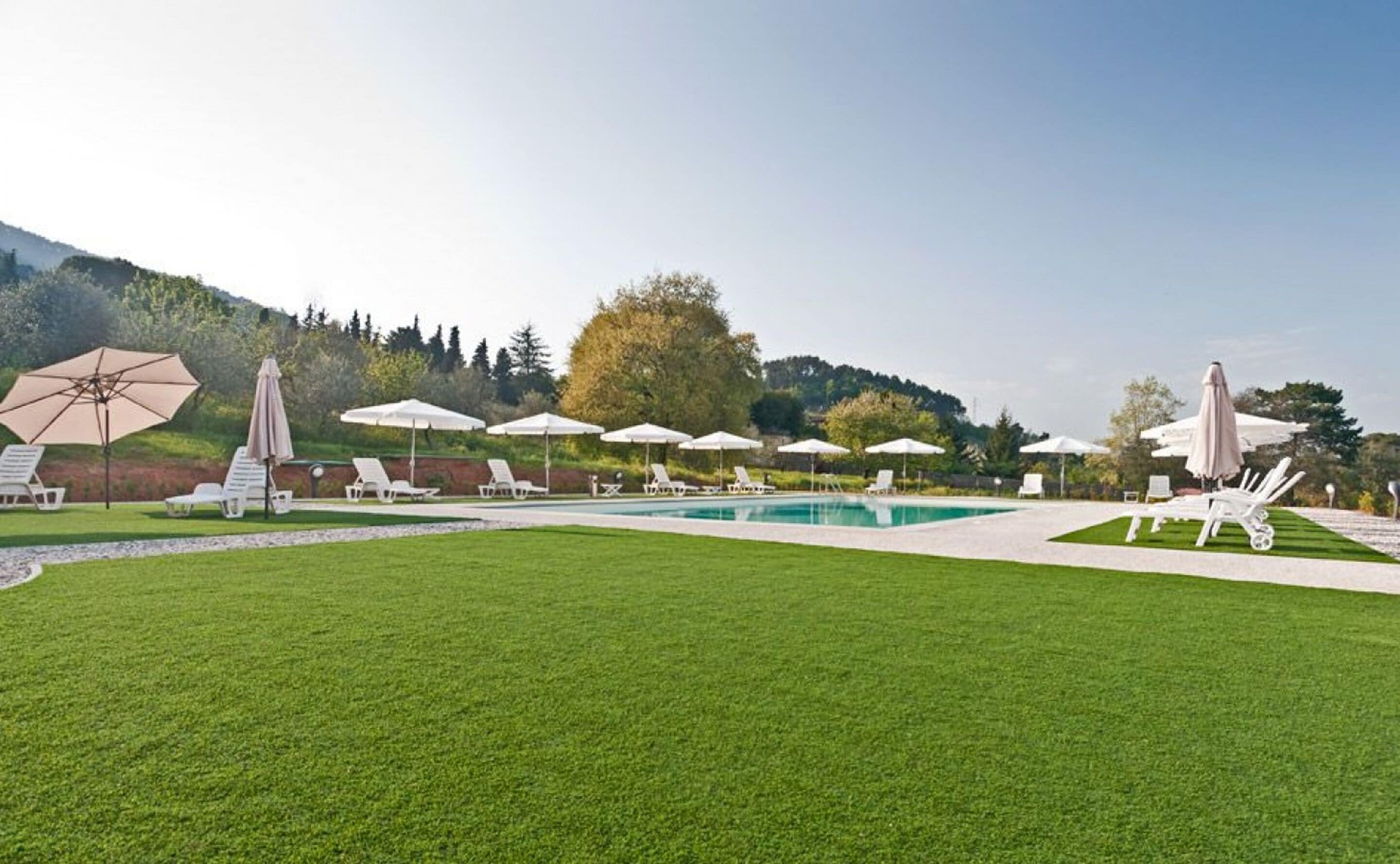 A pool area with white lounge chairs and umbrellas, surrounded by a well-manicured lawn and trees.