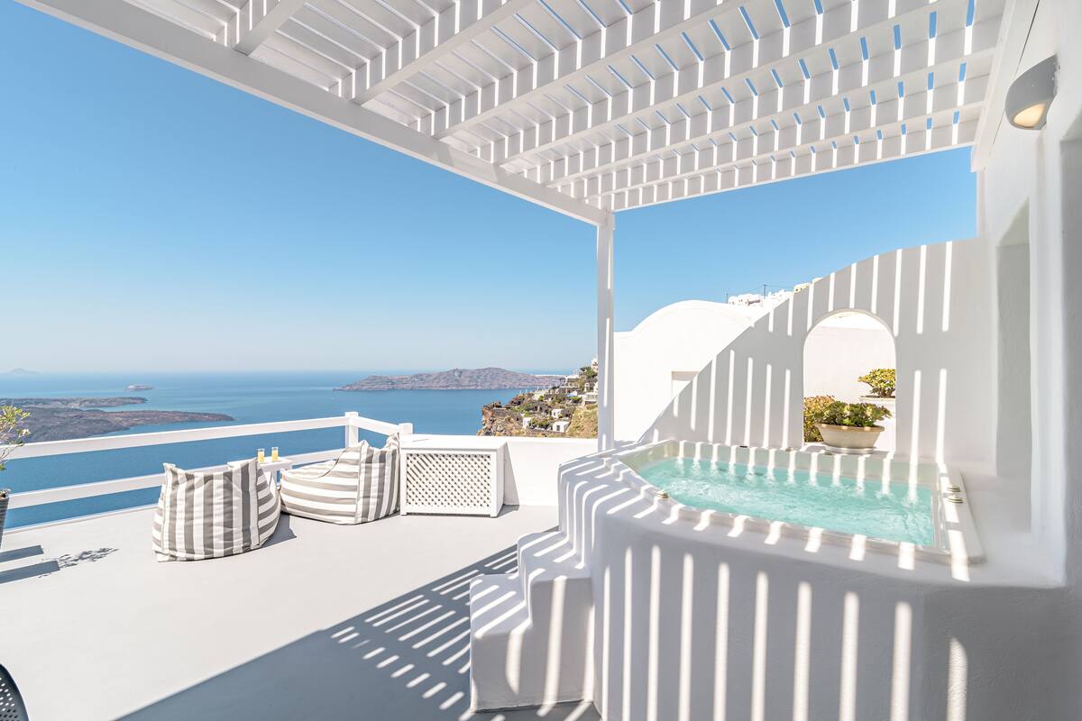 A white outdoor terrace with a pool, striped cushions, and a view of the sea.