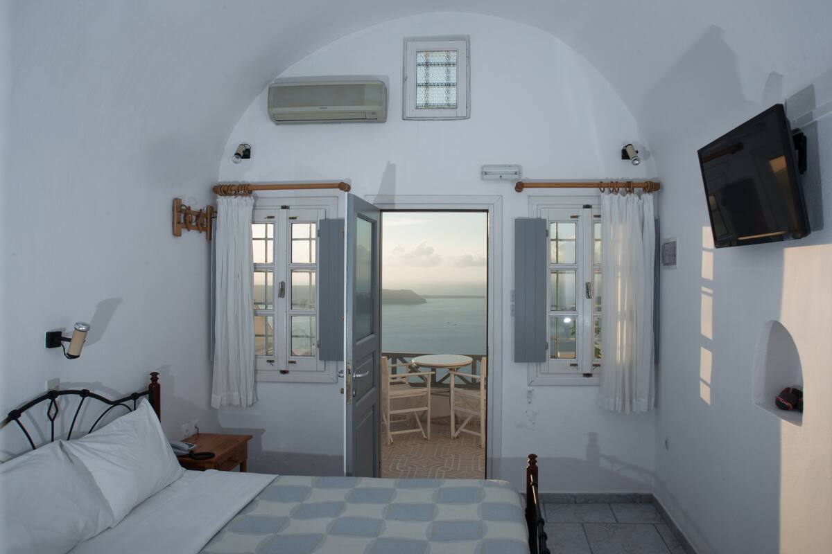 Un dormitorio con cama, mesita de noche, televisor montado en la pared y vista al mar a través de puertas abiertas.