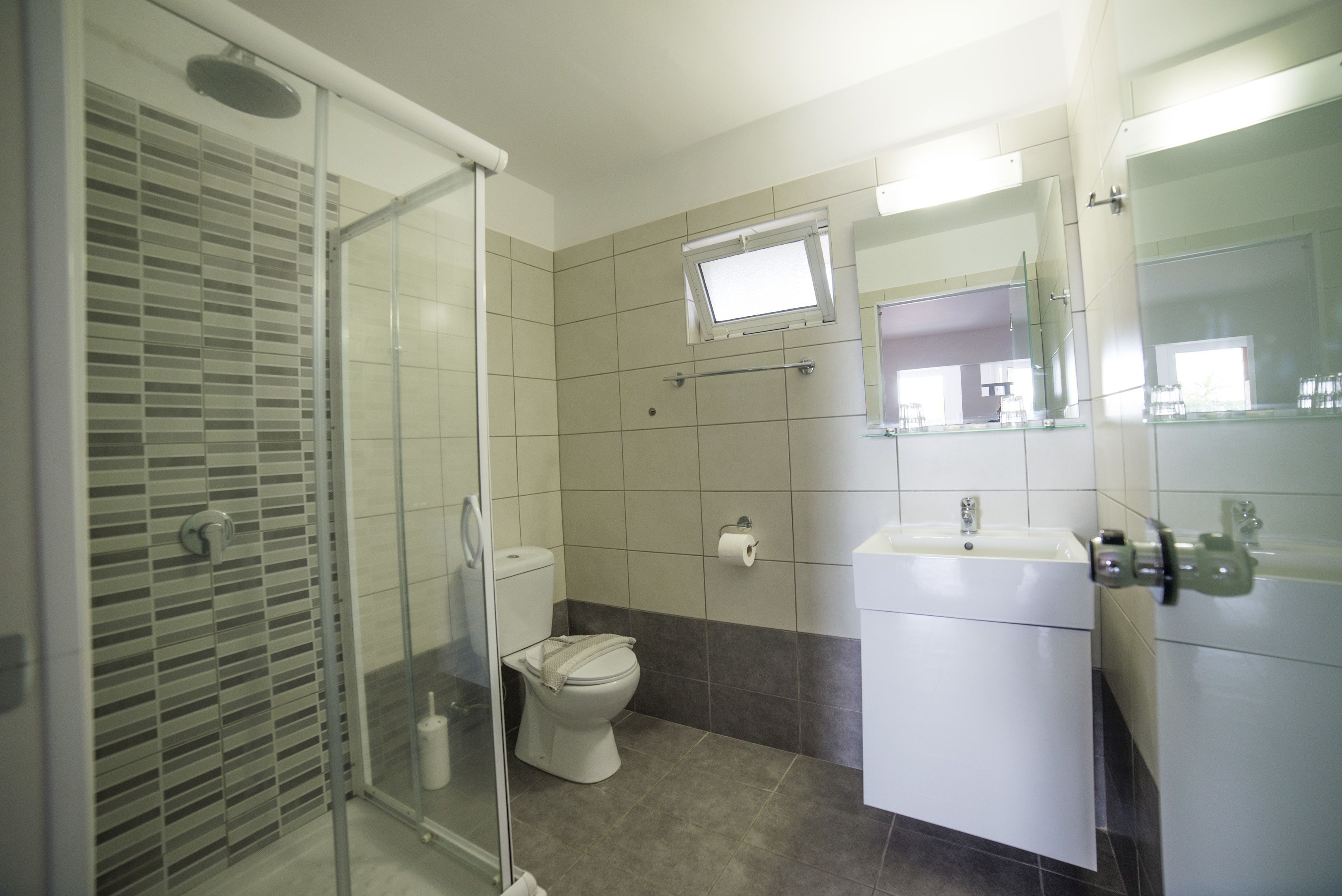 A bathroom with a glass shower enclosure, a toilet, a sink with a mirror, and a window above the toilet.