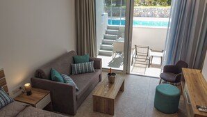 En moderne stue med sofa, sofabord og skammel. Champagne og vinglas står på bordet. Store vinduer giver udsigt til en pool og et udendørs siddeområde.
