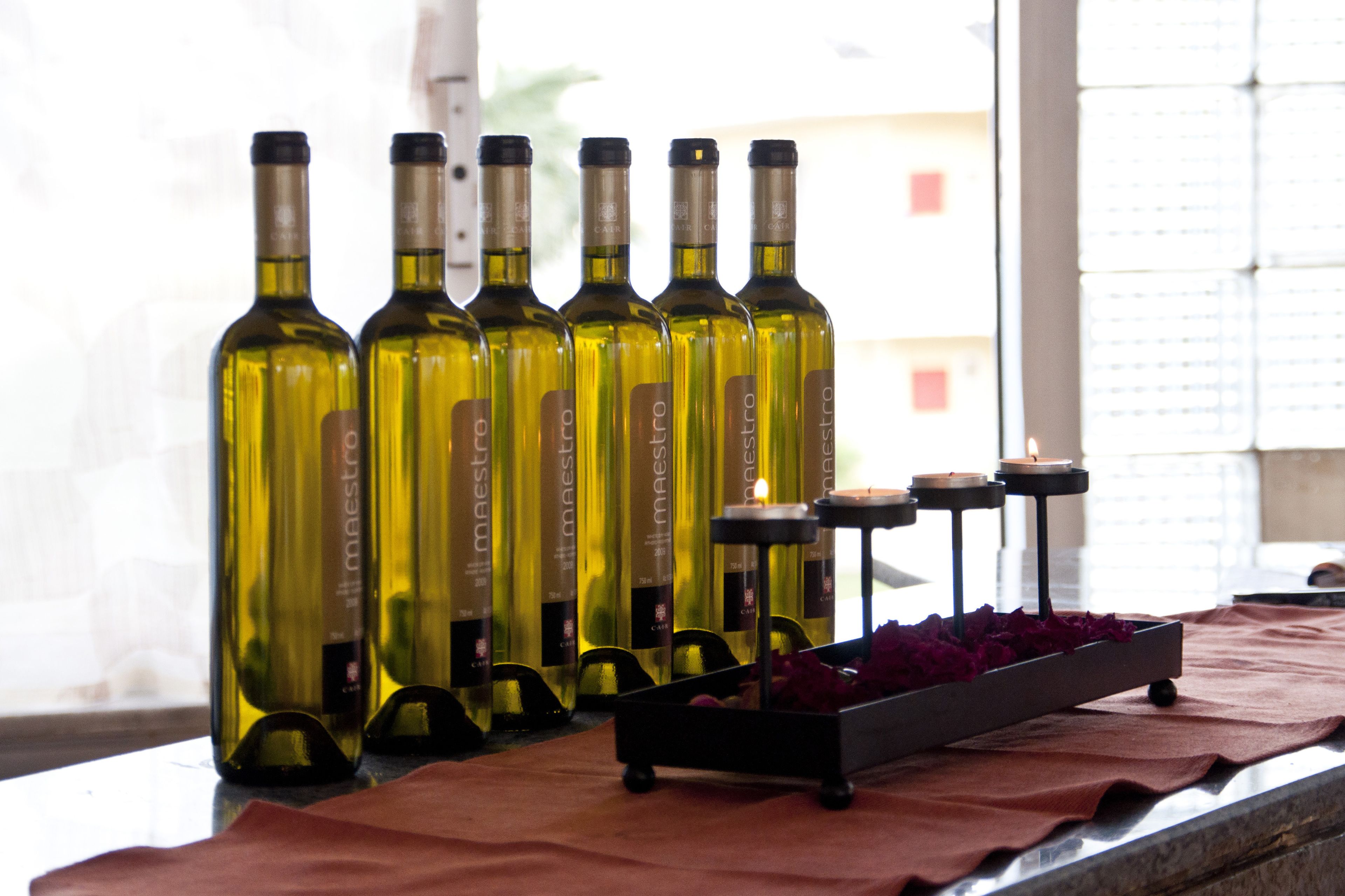 Una hilera de botellas de vino sobre una mesa con velas y una bandeja de pétalos de rosa.