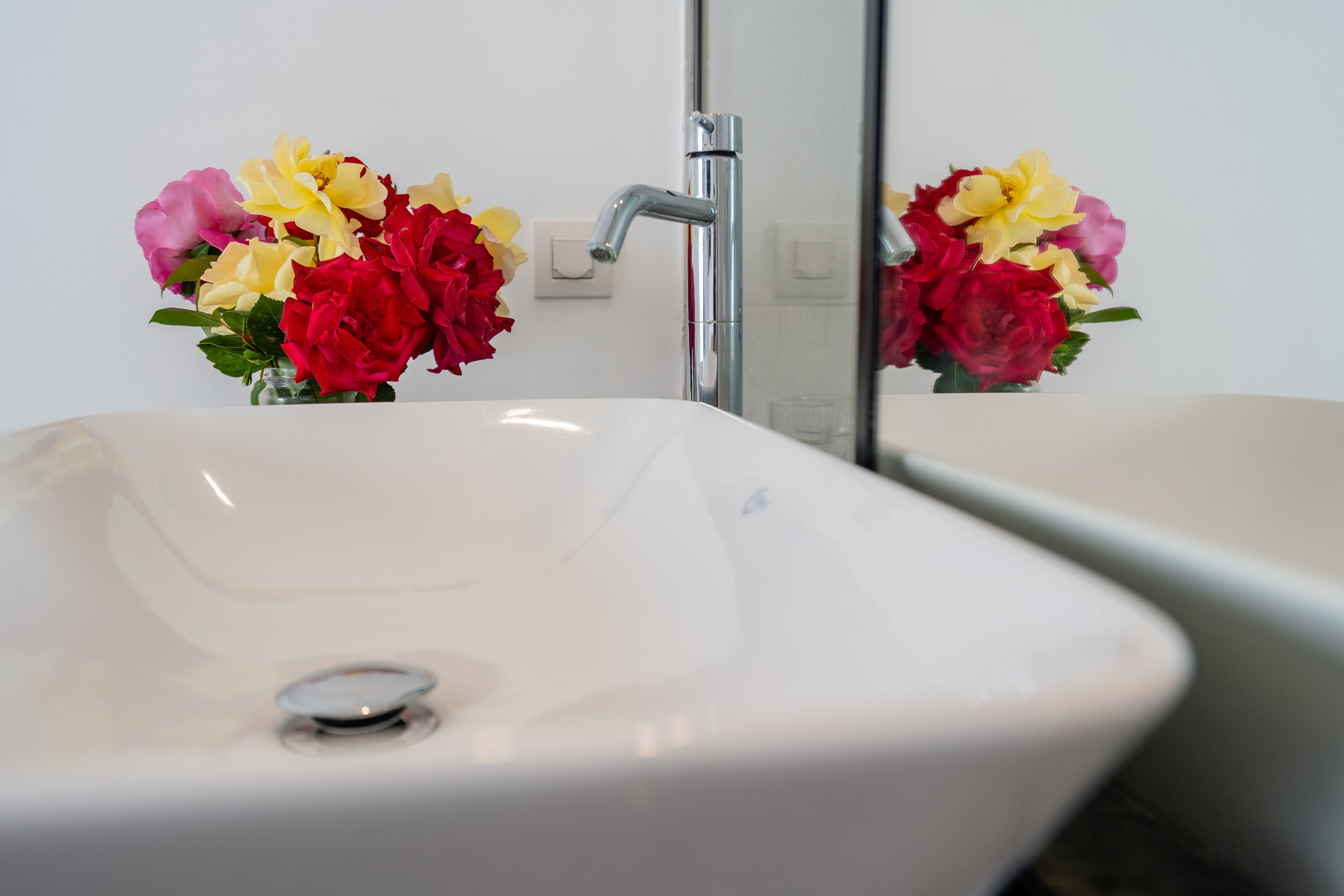 A bathroom with a white sink and a mirror reflecting a bouquet of flowers.