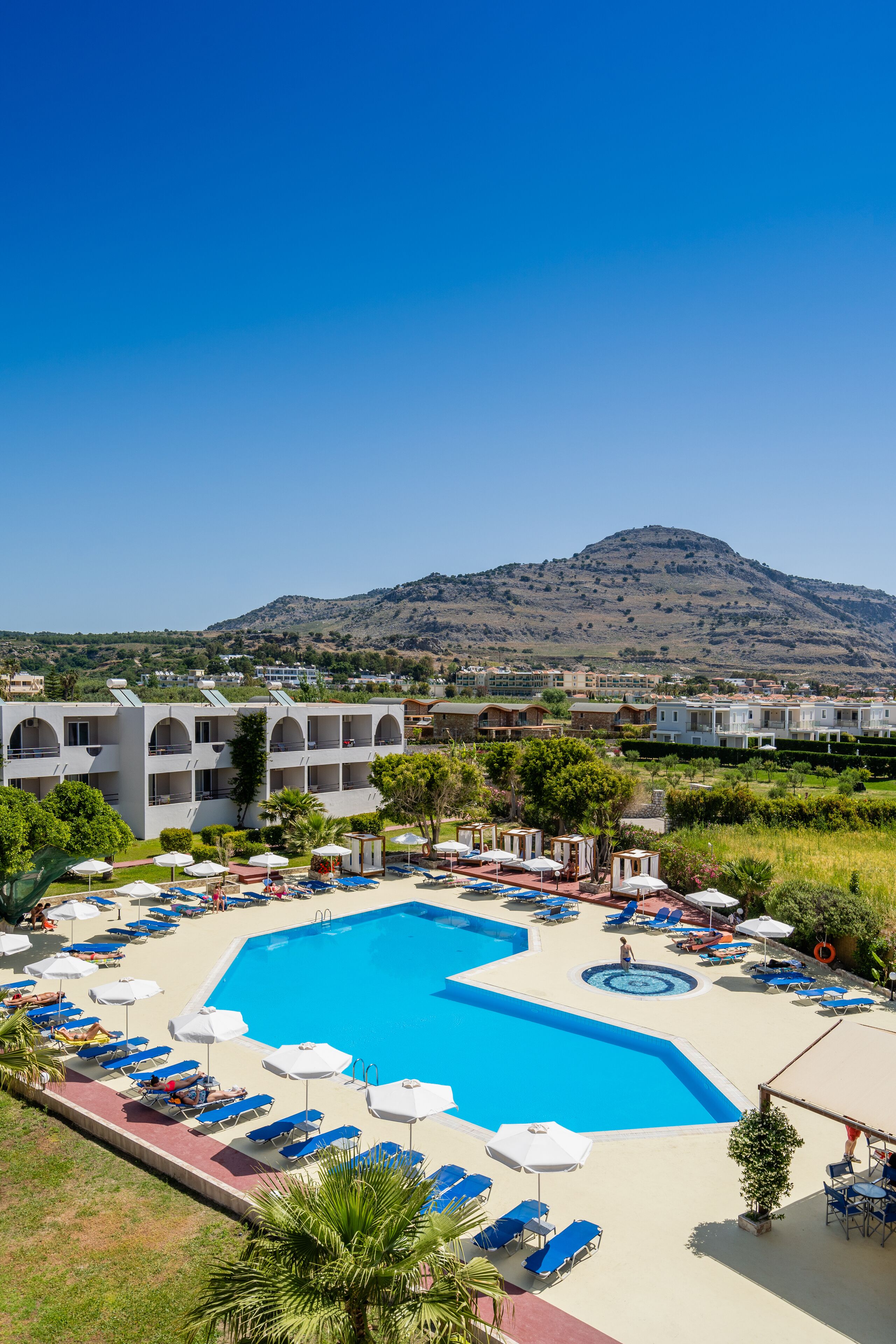 A resort with a large swimming pool, multiple lounge areas with umbrellas, and a view of a mountain in the background.