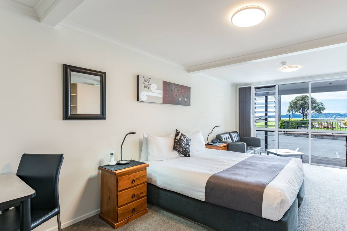 Habitación de hotel con cama, mesitas de noche, escritorio, una silla y un amplio ventanal con vistas al exterior.