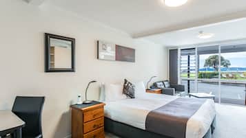 A hotel room with a bed, bedside tables, a desk, a chair, and a large window offering a view of the outdoors.
