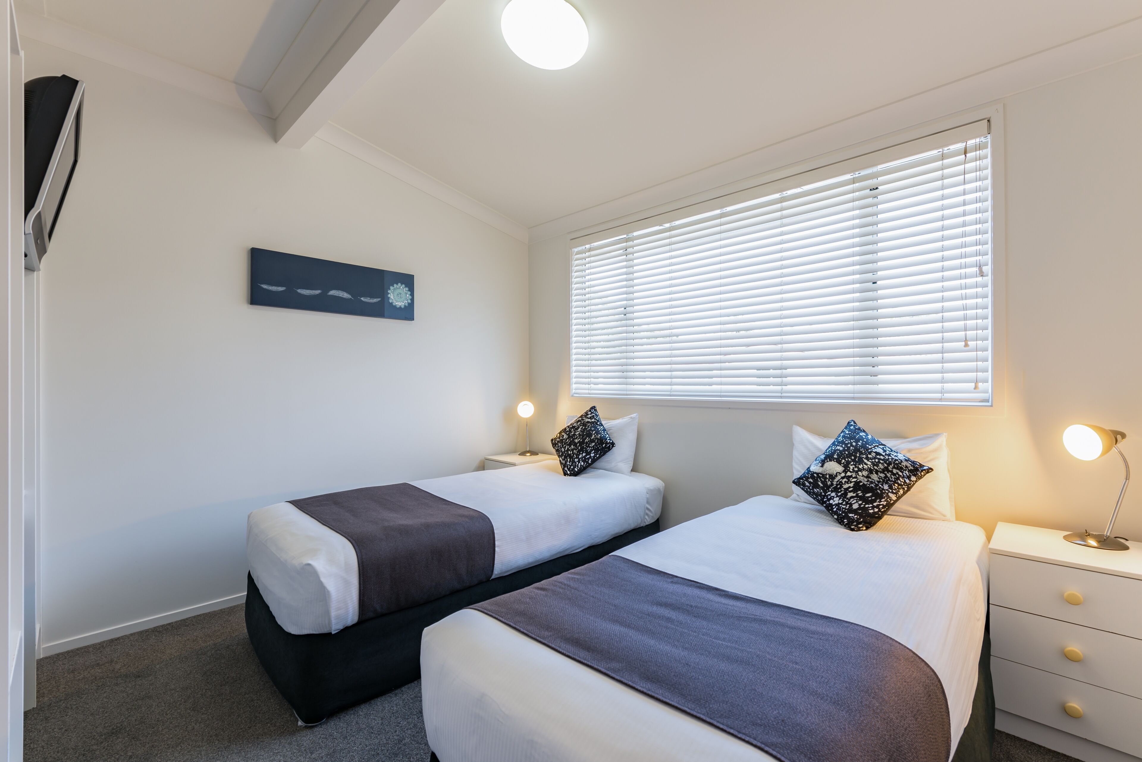 A hotel room with two beds, a nightstand with a lamp, a wall-mounted artwork, and a window with blinds.