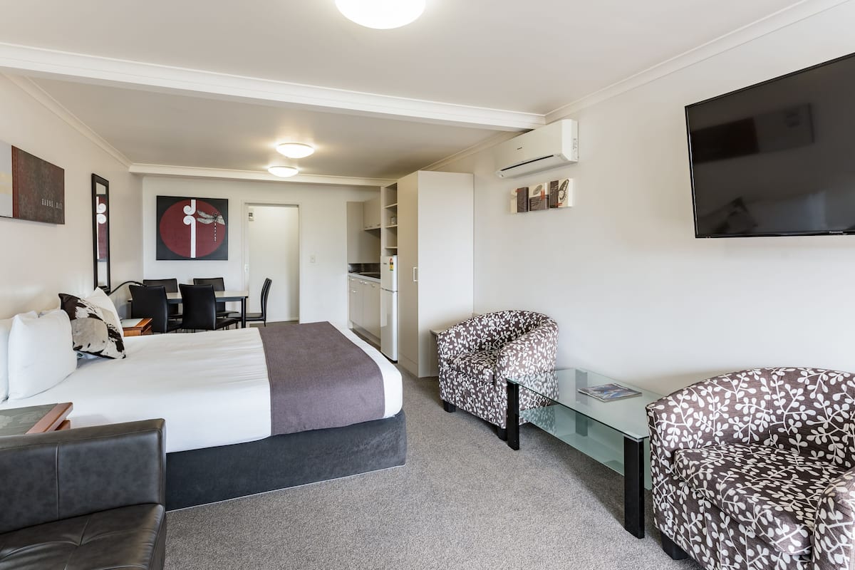 Una habitación de hotel moderna con cama, sofá, mesa de vidrio, silla, televisión y kitchenette.