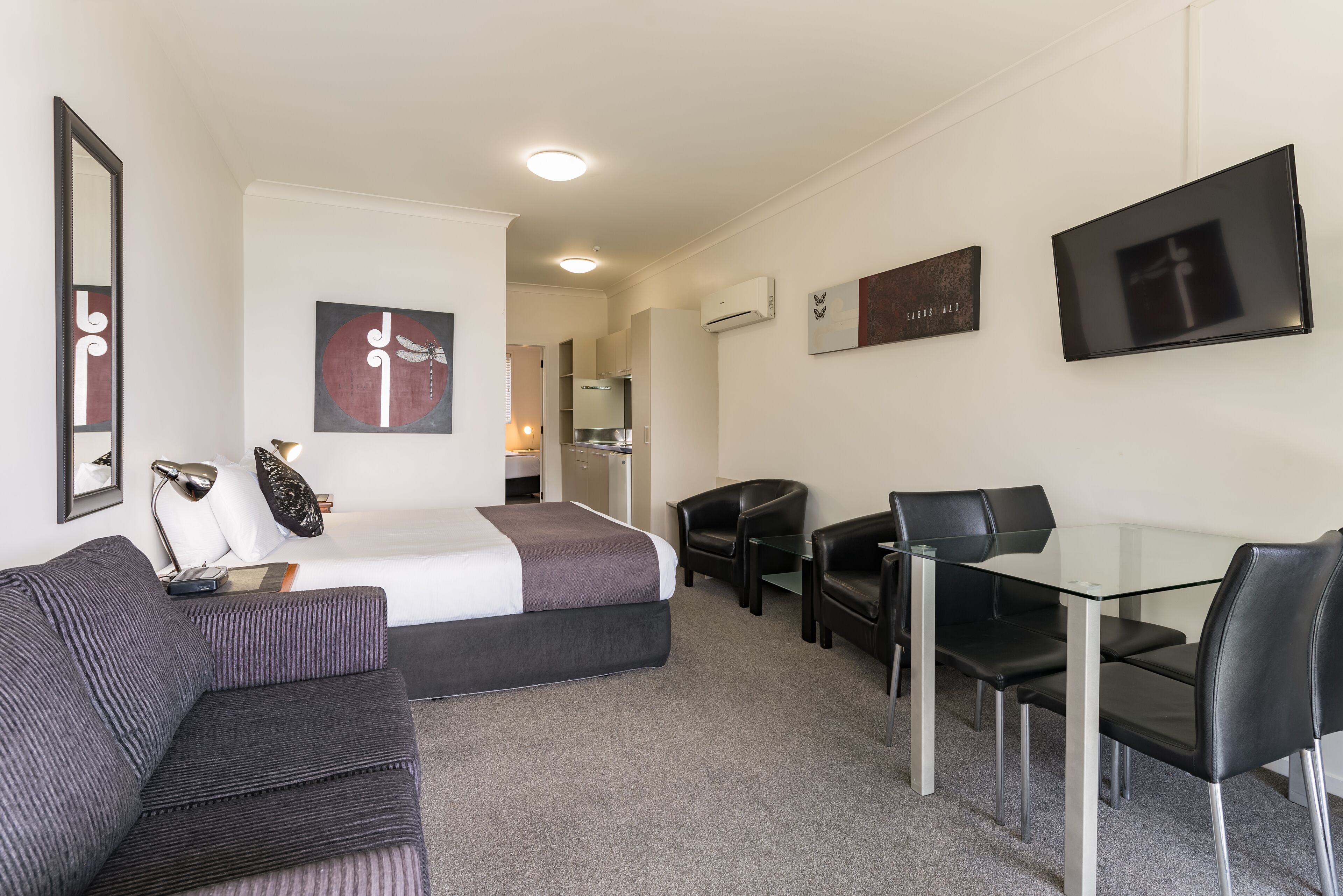 A hotel room with a bed, a sofa, a desk with chairs, a TV, and artwork on the walls.