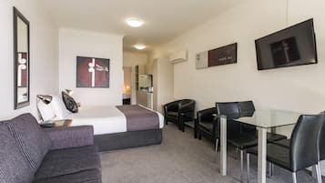 A hotel room with a bed, a sofa, a desk with chairs, a TV, and artwork on the walls.