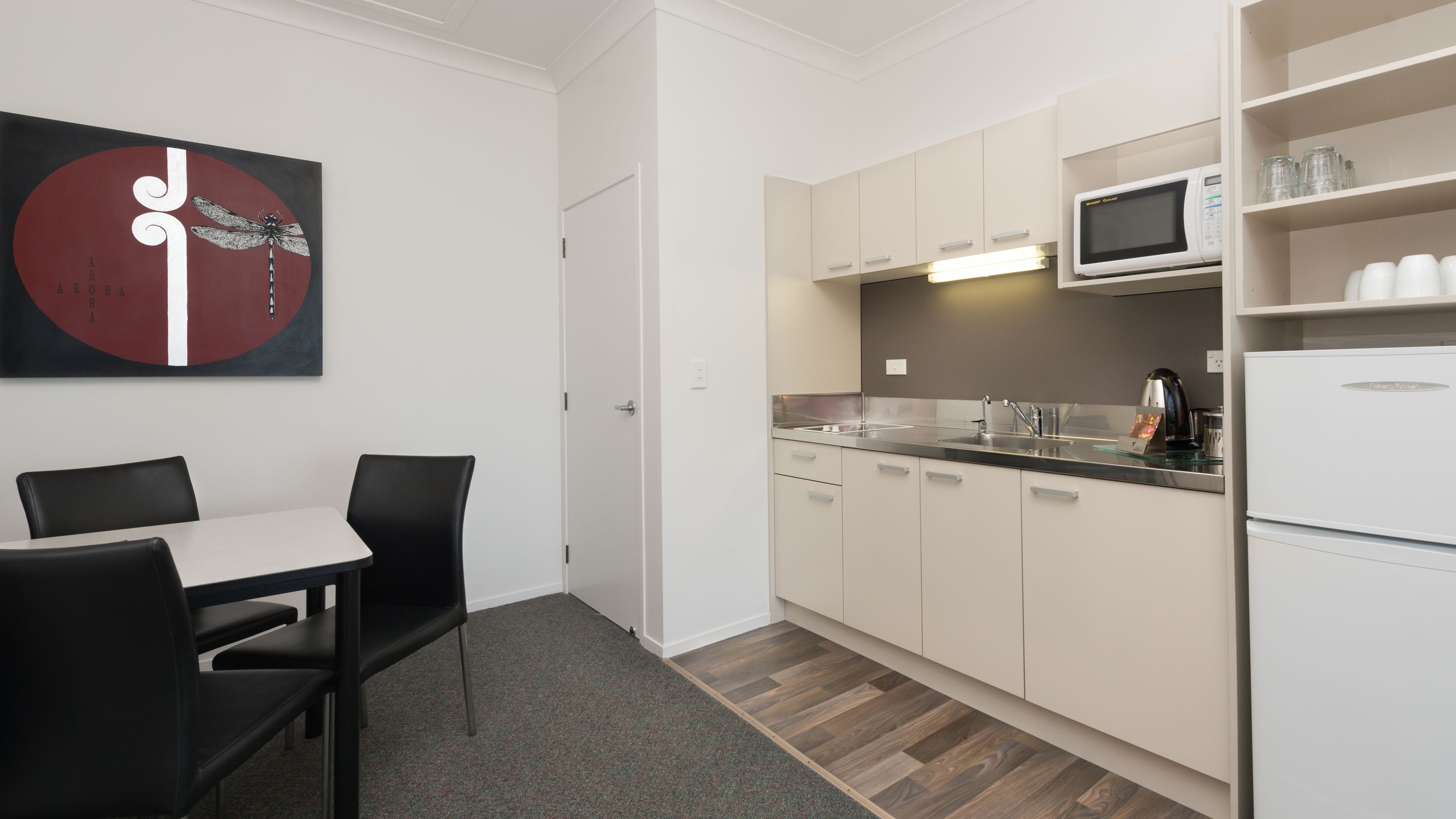 A modern kitchen with white cabinets, a microwave, and a refrigerator. There is a dining table with chairs and a wall-mounted artwork.