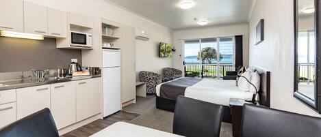 A compact hotel room with a kitchenette, a bed with a view, a small seating area, and a balcony with a view.