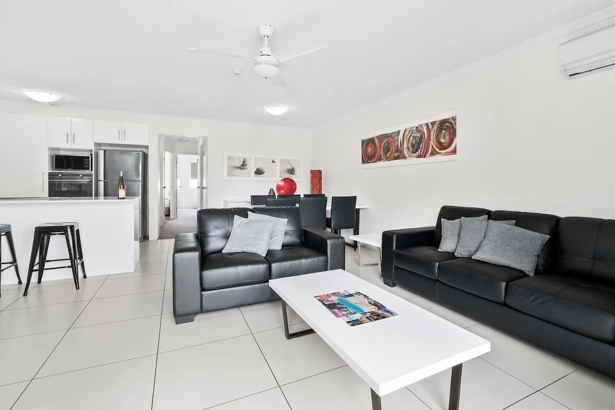 A modern living room with two black leather sofas, a white coffee table, and a kitchen area with white cabinets and appliances.