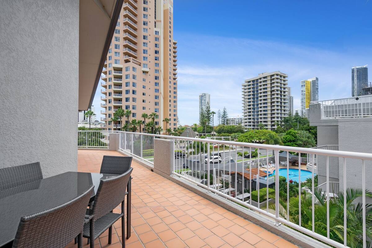 A balcony with a view of a swimming pool, high-rise buildings, and a fenced area with greenery.