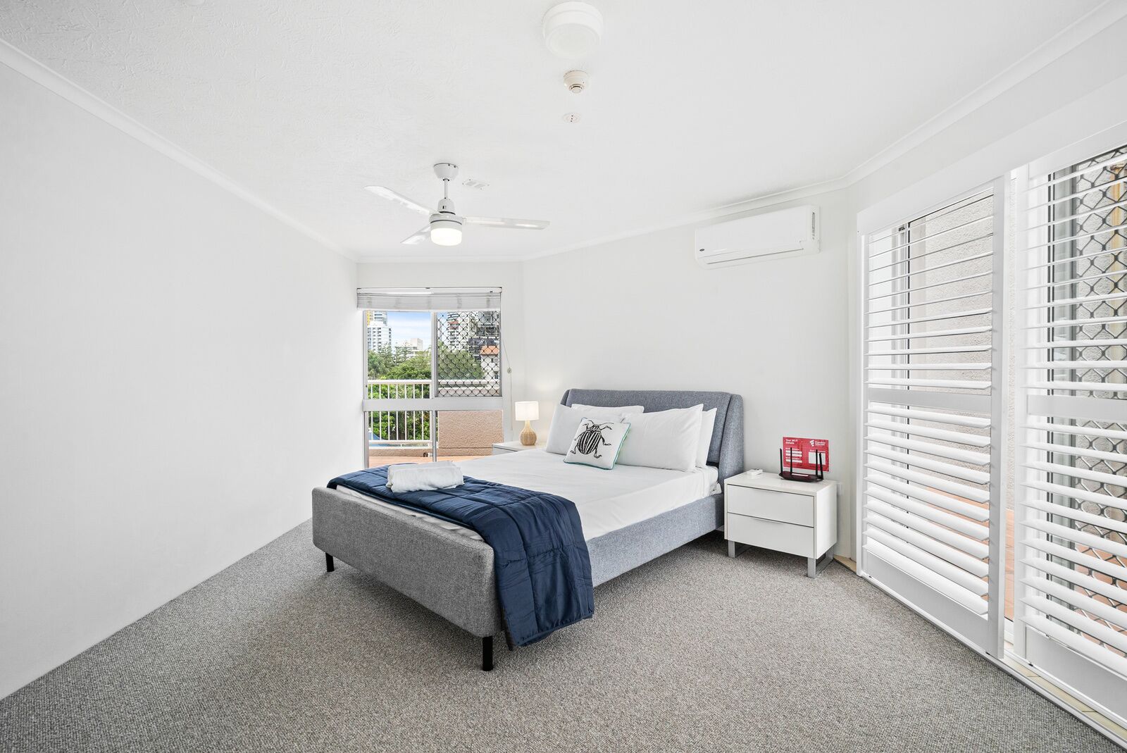 Une chambre à coucher comprenant un lit, une table de chevet, une commode, un ventilateur de plafond et une fenêtre avec des stores.