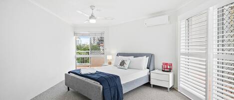 Une chambre à coucher comprenant un lit, une table de chevet, une commode, un ventilateur de plafond et une fenêtre avec des stores.