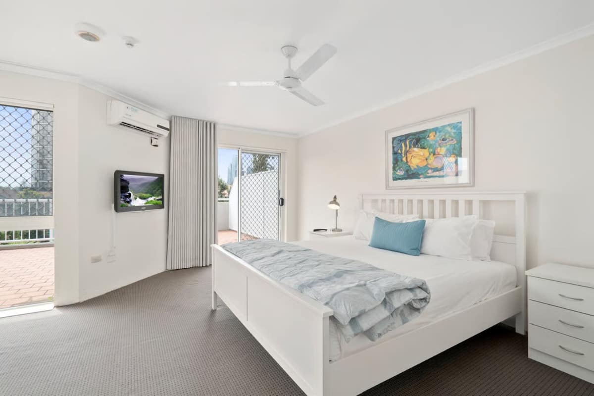 A bedroom with a white bed, a ceiling fan, an air conditioning unit, a TV, a painting, and a view of the outdoors.