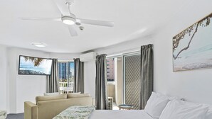 Habitación de hotel con cama, sofá, televisión, ventilador de techo y ventana con vista a edificios.