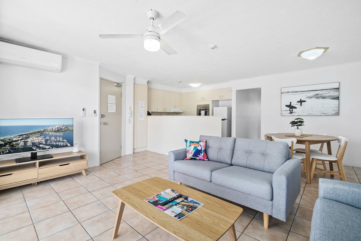 A modern living room with a grey sofa, a wooden coffee table, a flat-screen TV, a ceiling fan, and a kitchen area visible through an open door.
