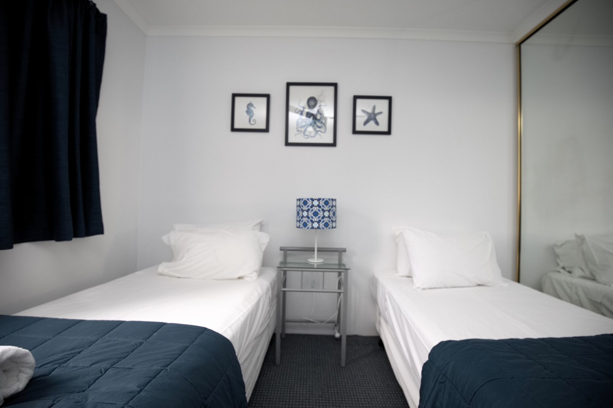 Une chambre d’hôtel avec deux lits simples, une petite table avec une lampe et trois tableaux encadrés accrochés au mur.