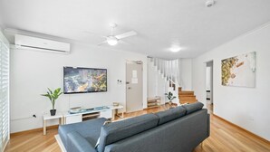 Una sala de estar moderna con un sofá gris, un televisor de pantalla plana, un ventilador de techo y una escalera que sube a otro nivel.
