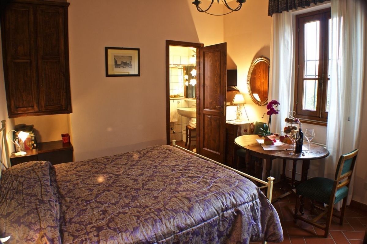 Un dormitorio con cama, mesita de noche, una pequeña mesa redonda con lámpara, una silla y una ventana con cortinas.