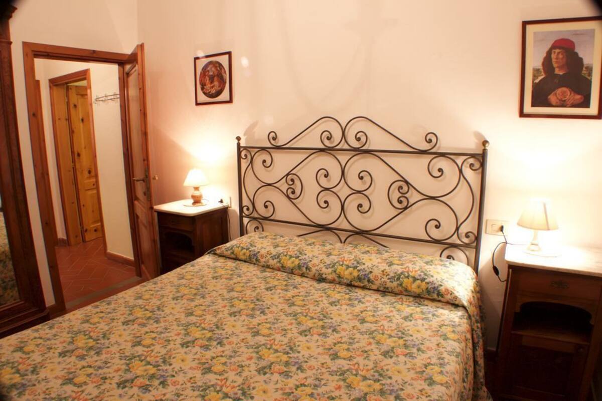 A bedroom with a floral bedspread, a metal headboard, two bedside tables with lamps, and a door leading to another room.