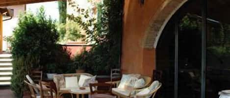 Un patio al aire libre con mesas y sillas de madera, rodeado de follaje y un muro de ladrillo.